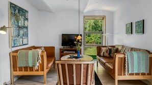 Una sala moderna con dos sofás, una mesa de centro y un televisor. Una ventana grande ofrece vistas a zonas verdes.