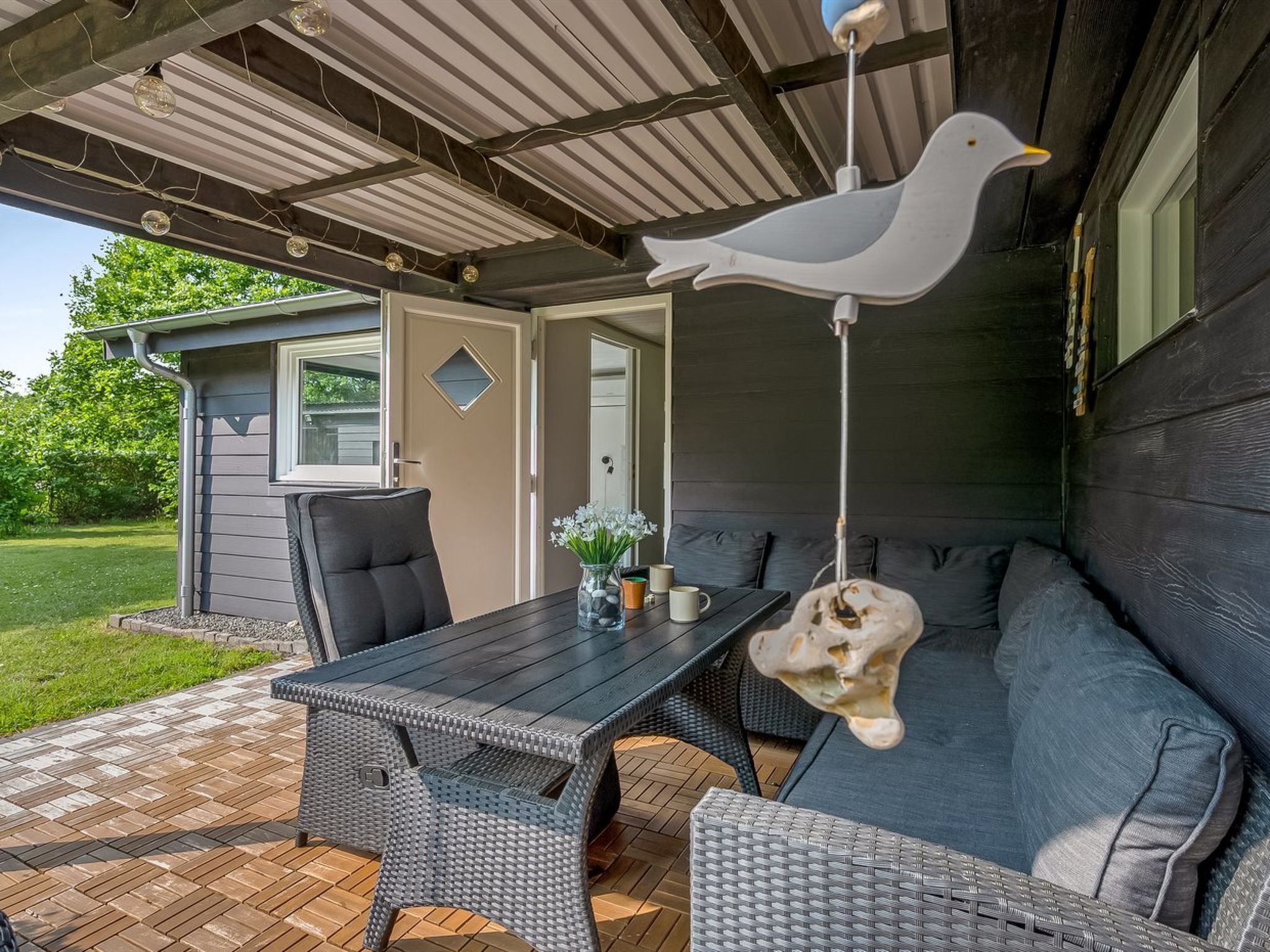 An outdoor patio with a dark-colored table and wicker chairs, a hanging bird decoration, and a small shed in the background.
