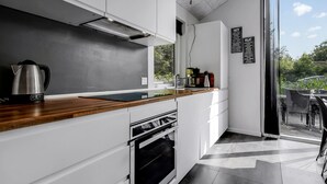 A modern kitchen with white cabinetry, a wooden countertop, a kettle, a coffee machine, and a view of the outdoors through a glass door.