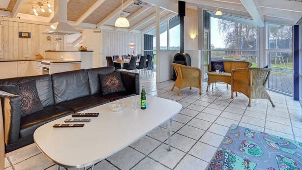 A spacious living area with a sofa, a coffee table, and wicker chairs. There is a kitchen in the background with a dining area.