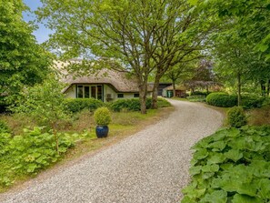 Un chemin de gravier mène à un bâtiment au toit de chaume, entouré d’une végétation luxuriante et d’un jardin bien entretenu.