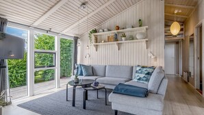 A modern living room with a grey sectional sofa, a coffee table, and a TV on a stand. There are shelves with decorative items and a large window with a view of greenery. - "Asko" - 350m from the sea by Interhome (Ebeltoft)