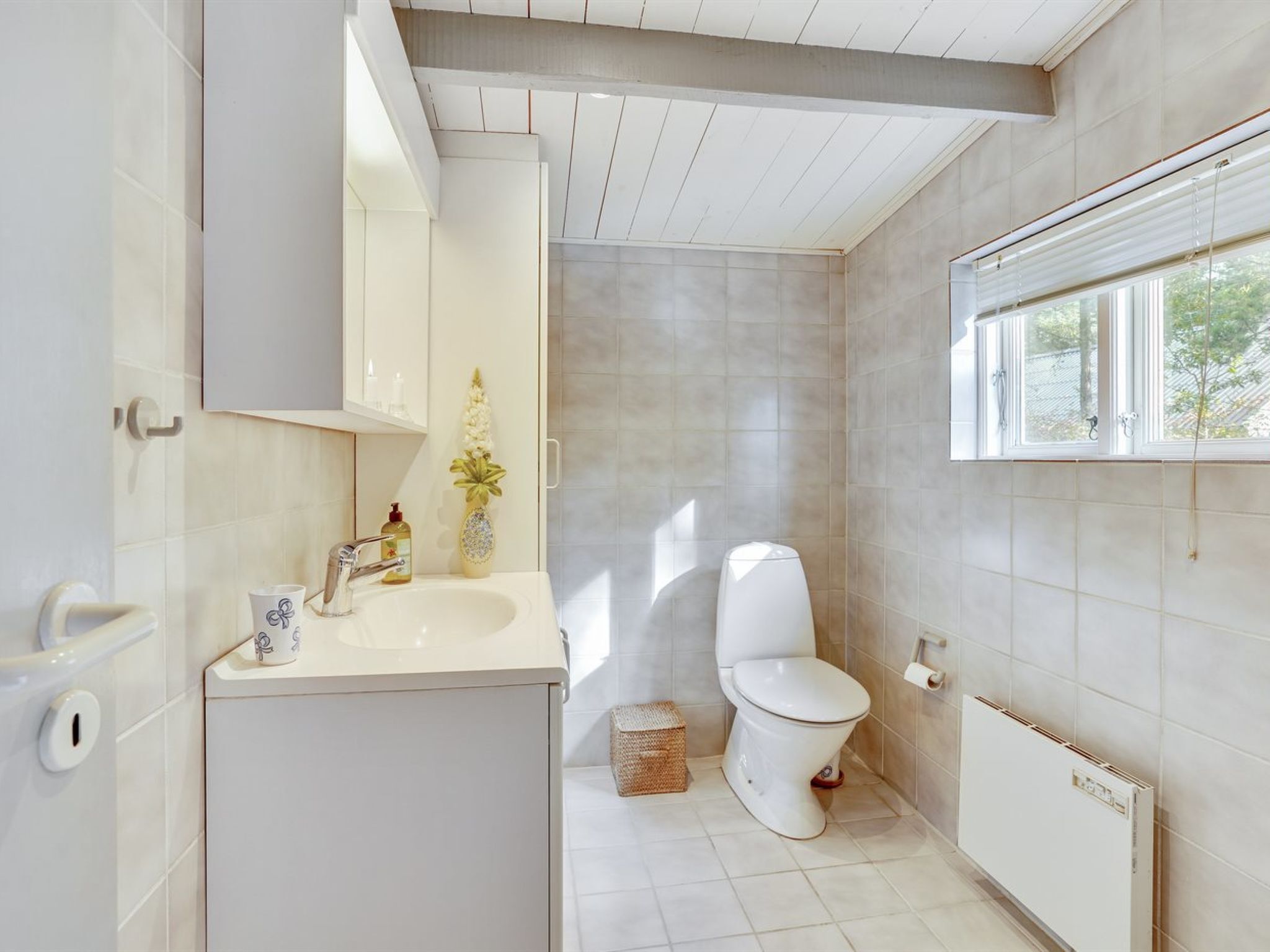 Un baño moderno con un lavabo, inodoro y radiador blancos. Hay una ventana que proporciona luz natural y un jarrón decorativo sobre el lavabo.