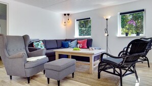 A modern living room with a grey sofa, ottoman, and black wicker chairs. There is a wooden coffee table with decorative items and a view of greenery through the windows. - "Tannie" - 300m from the sea by Interhome (Strøby)