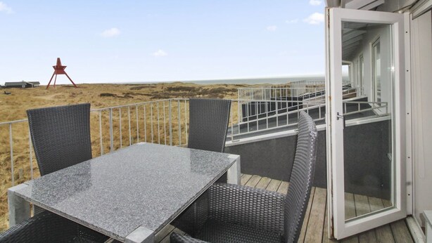 Una terraza con mesa y sillas, con vista a una playa y una estructura roja a lo lejos.