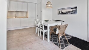 Una cocina moderna con armarios blancos y encimera de madera, un comedor con mesa blanca y sillas de mimbre, y una alfombra blanco y negro en el suelo.