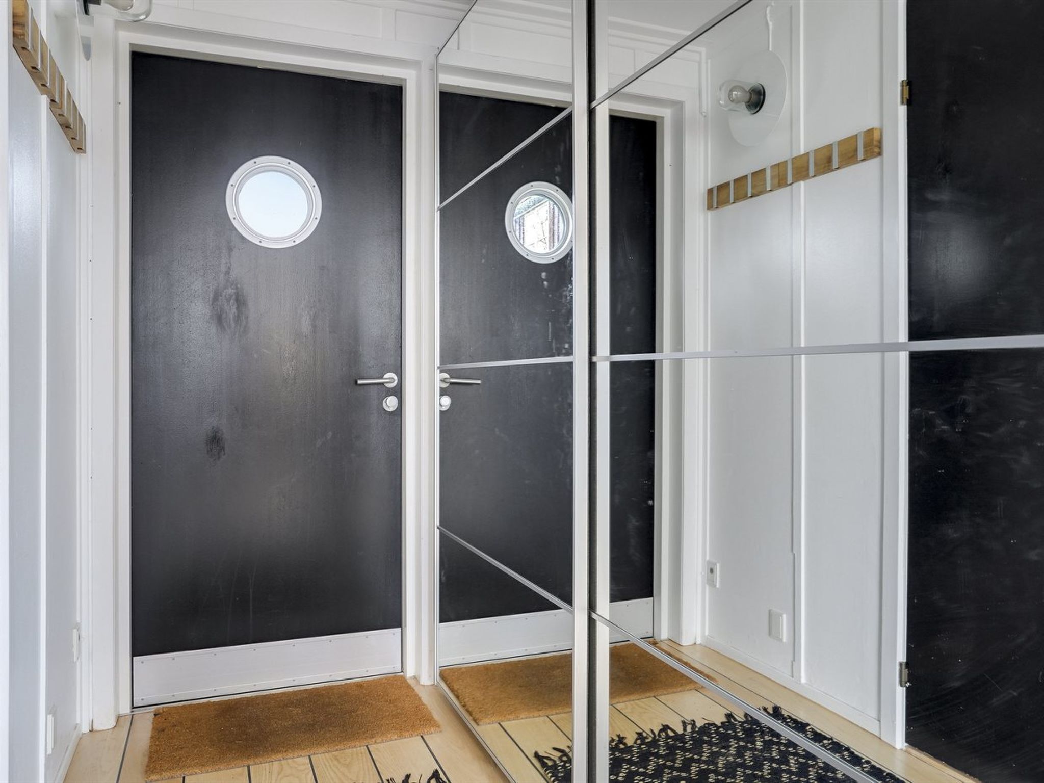 Une pièce avec une porte noire, une porte blanche et une porte en verre dotée d’une poignée en métal. Le sol est en bois et il y a un paillasson devant la porte.