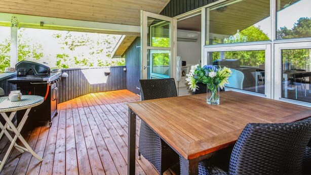 Een terras buiten met een houten terras, een barbecue, rieten meubilair en grote ramen met uitzicht op groen.
