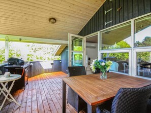 Une terrasse extérieure dotée d’une plateforme en bois, d’un barbecue, de meubles en osier et de grandes fenêtres offrant une vue sur la verdure.