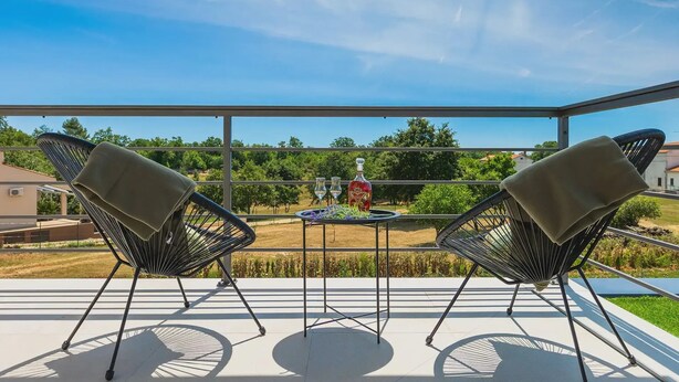 A balcony with two chairs, a table with a bottle and glasses, and a view of a garden and buildings. - Artsi in Svetvin Enat (Svetvincenat)