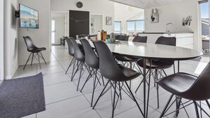 A modern dining area with a large round table and black chairs, a kitchen in the background, and a view of the outdoors.