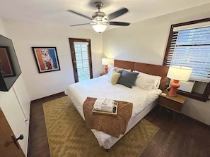 A hotel room with a bed, a ceiling fan, a window with blinds, a bedside lamp, a framed picture on the wall, and a small table with a tray.