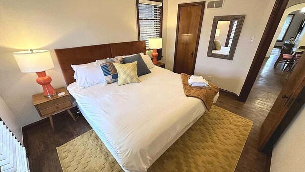 A hotel room with a large bed, two bedside tables, a lamp, a mirror, and a door leading to another room.