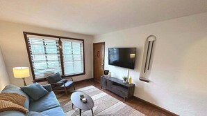 A living room with a sofa, chairs, a coffee table, a TV, and a window with blinds.