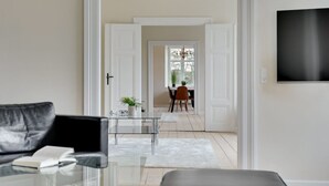 Un salon moderne avec une table basse en verre, un canapé en cuir et un pouf noir. On aperçoit un coin repas à travers une porte ouverte, avec une table à manger, des chaises et un lustre. Un téléviseur à écran plat est fixé au mur.