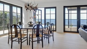 Ein moderner Essbereich mit großem Tisch und Stühlen, einer außergewöhnlichen Wandleuchter aus Geweihen und bodengroßen Fenstern mit Meerblick.
