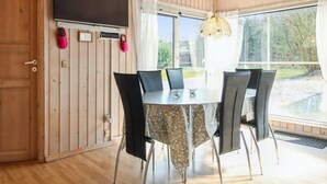 Une pièce aux murs lambrissés, comprenant une table à manger, des chaises, un téléviseur fixé au mur et une grande fenêtre donnant sur un paysage verdoyant.