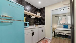 Cocina compacta con un refrigerador turquesa, un microondas y un lavamanos. Hay una cama con un almoháron azul y una ventana con persianas.
