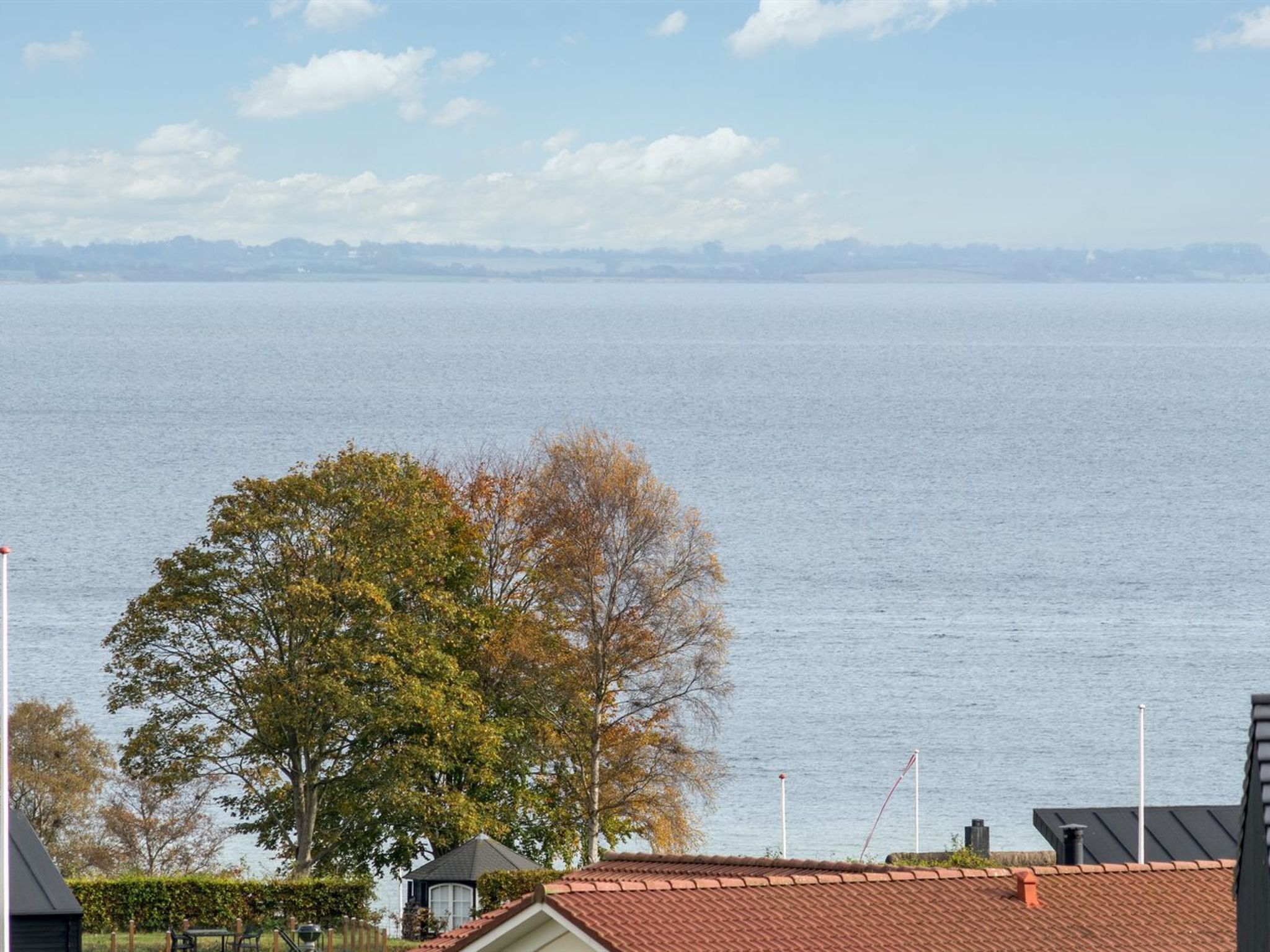 A coastal view with a house and a large tree in the foreground, a clear sky, and calm sea.