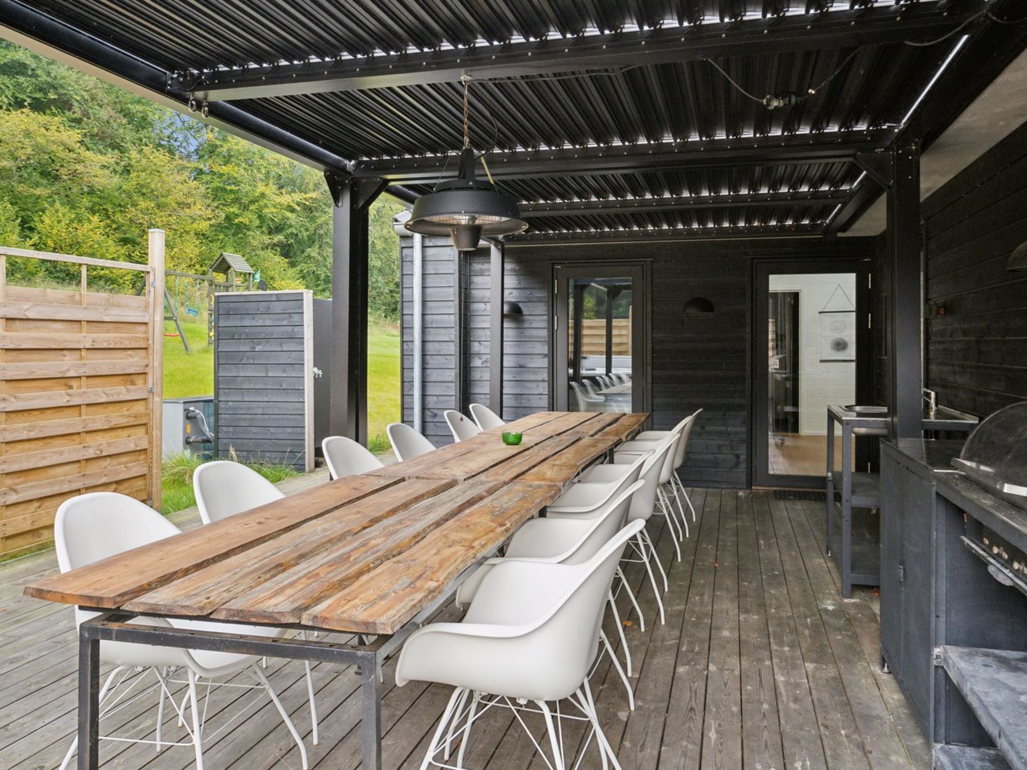 Et udendørs spiseområde med et langt træbord og hvide stole, grillstation og et overdækket siddeområde med ståltag.