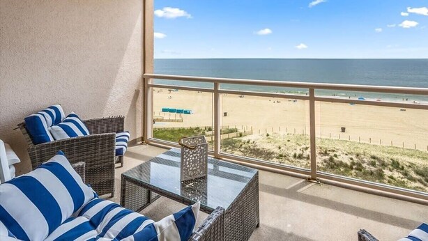 Een balkon met rieten meubilair en een glazen tafel, met uitzicht op een strand onder een heldere lucht.