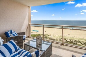A balcony with wicker furniture and a glass table, overlooking a beach with a clear sky. - Oceanfront Condo with Pool and Close to Everything (Ocean City)