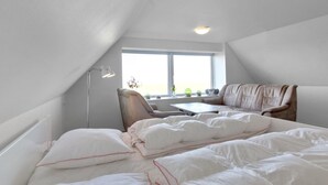 Une chambre avec un plafond en pente, un lit avec une literie blanche, un bureau, une chaise et une fenêtre donnant sur un beau paysage.