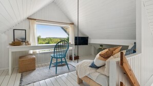 A bedroom with a sloped ceiling, a bed, a desk, a chair, and a window overlooking greenery.