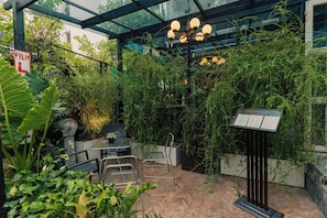 A greenhouse with a brick floor, metal chairs, a table, and a signboard.