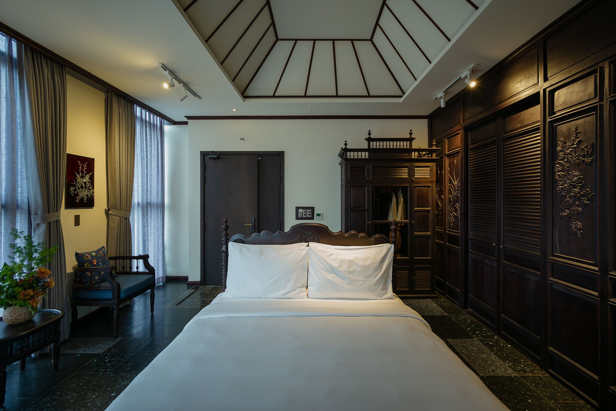 Una habitación de hotel con una cama grande, un armario de madera, un área de descanso con una silla y una mesita, y un ventanal con cortinas.