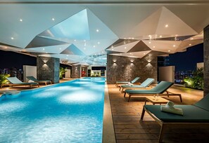 Un área de piscina interior moderna con diseños geométricos en el techo, sillas de descanso y vistas de la ciudad de noche.