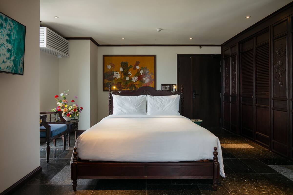 A hotel room with a large bed, a desk with a chair, a vase of flowers, and a painting on the wall.