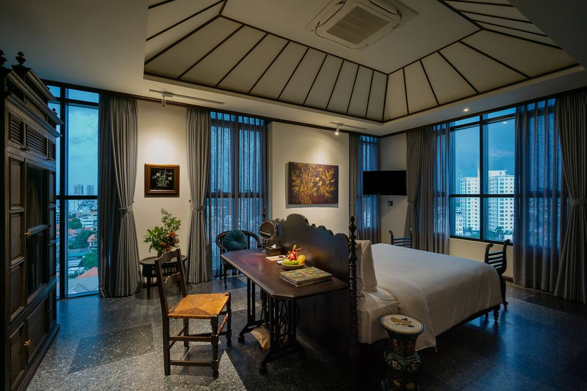 A hotel room with a large bed, a dining table with chairs, a small table with a plant, and a painting on the wall.