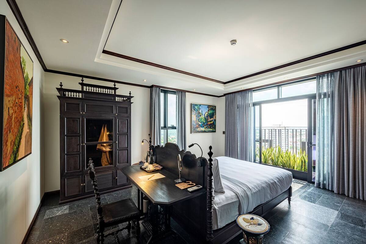 A bedroom with a large bed, a desk with a chair, a dark wooden cabinet, and a large window with curtains.