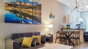 A modern living room with a sofa, a dining table with chairs, a kitchen area, and a large landscape painting on the wall. - The Stay at Steuben, Fire Pit, Brand new furniture (Springdale)
