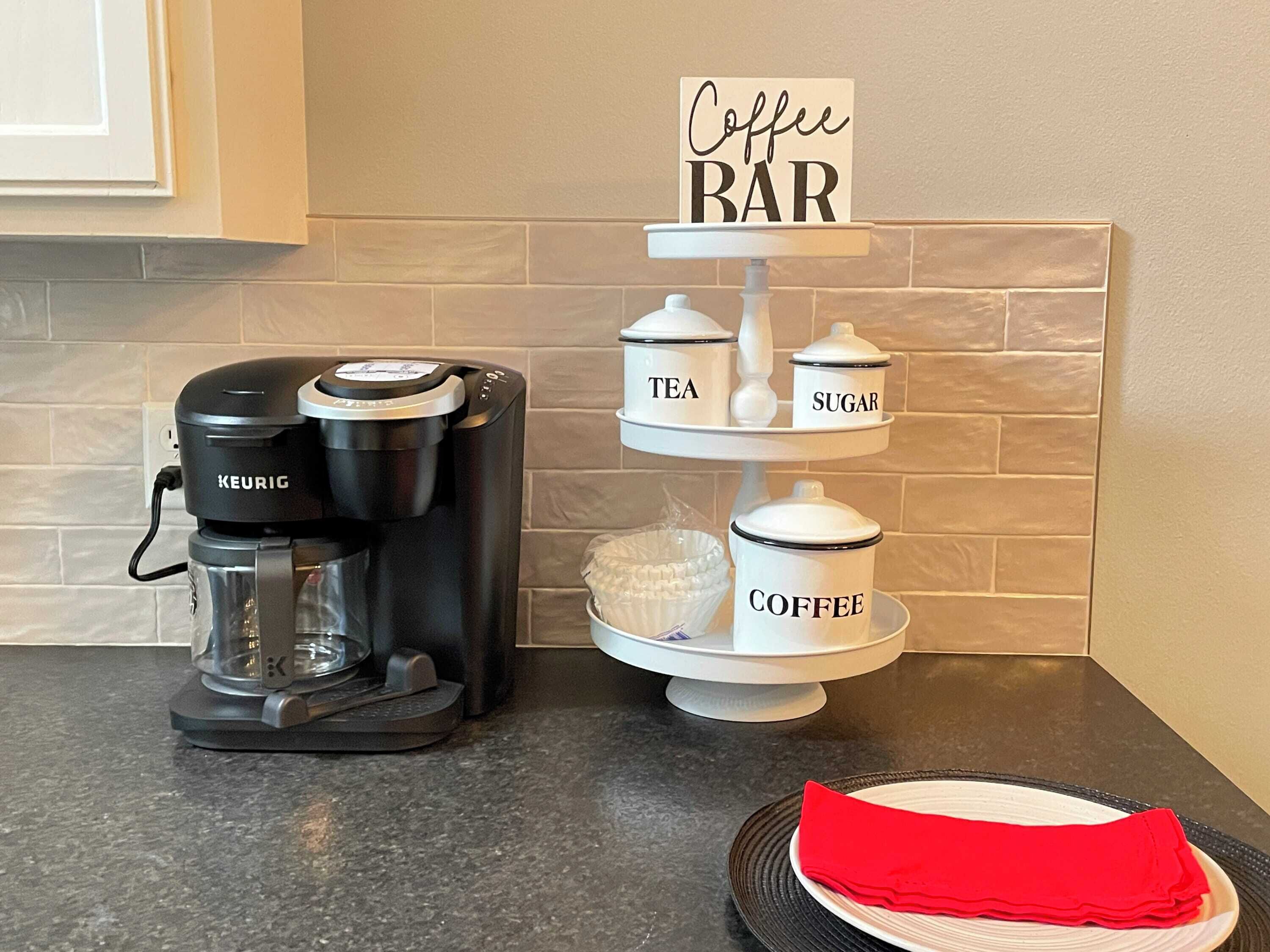 Uma cafeteira, um suporte escalonado com recipientes etiquetados para chá, açúcar e café, e um guardanapo vermelho sobre um prato.