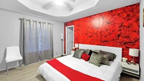Un dormitorio con una cama, ventilador de techo, pared con estampado floral rojo, una silla blanca y una puerta.
