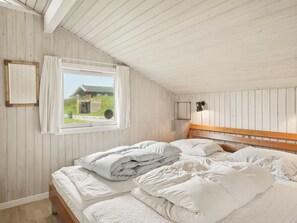 Un dormitorio con techo de madera, una cama con ropa de cama blanca, una ventana con cortinas y vistas a una casa exterior.