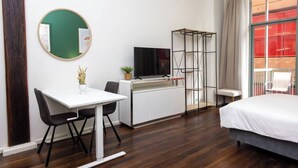 Een hotelkamer met een wit bureau, een televisie, een bed, een balkon met een rode reling en een groot raam.