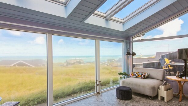 A room with large glass doors leading to a beach view, a sofa with cushions, a small round ottoman, and a table with a lamp.