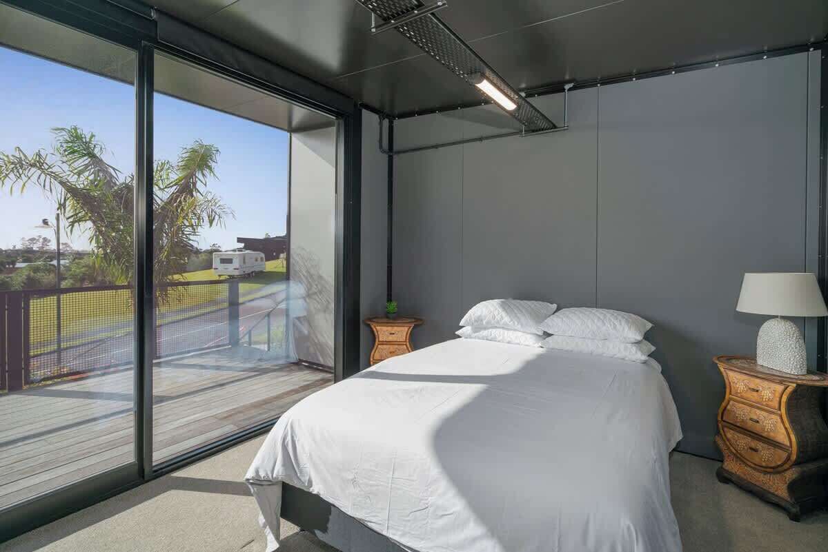 A bedroom with a large window, a bed, a nightstand, and a view of palm trees.