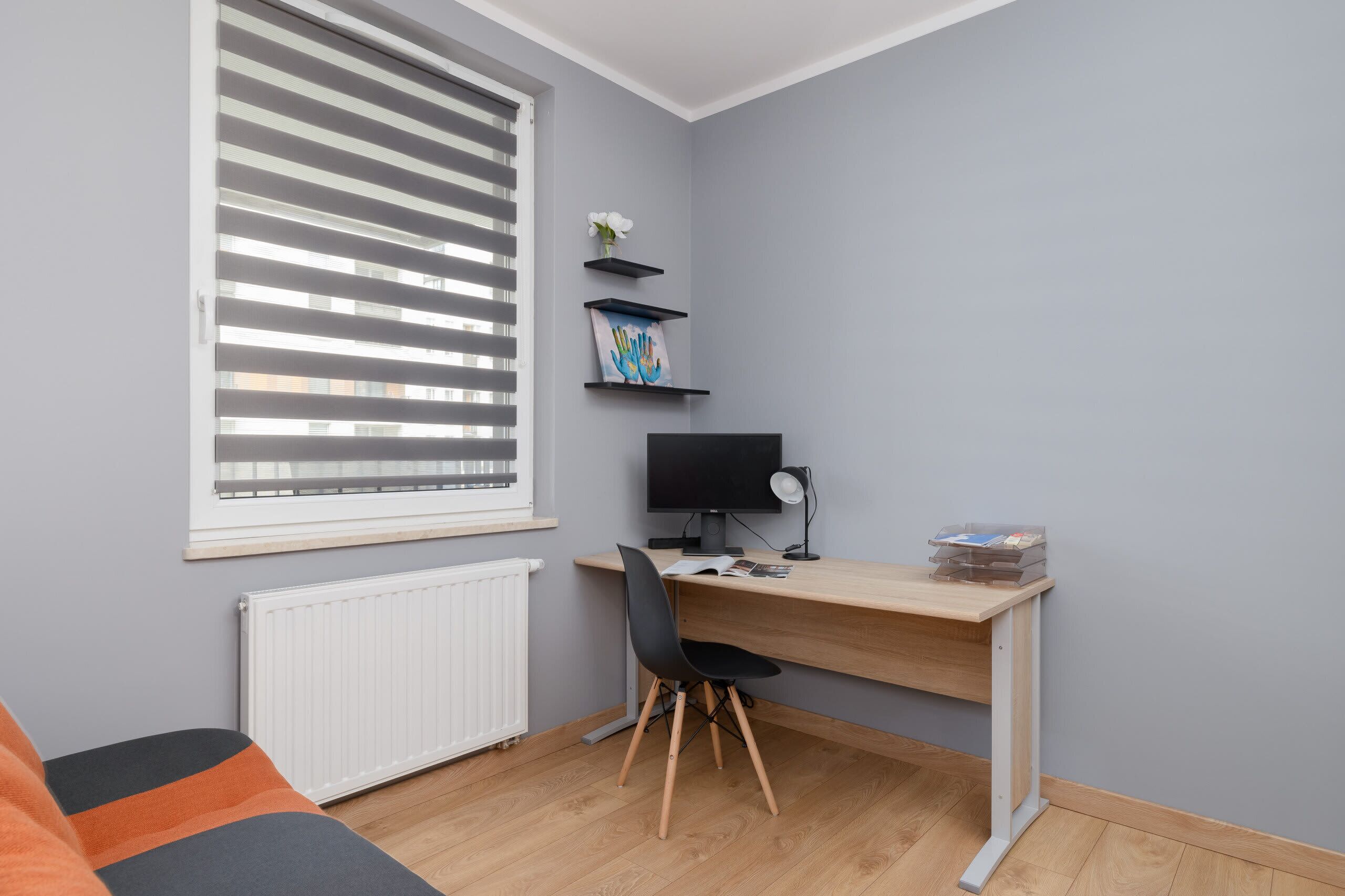 A room with a wooden desk, a black chair, a white radiator, a window with blinds, and a grey wall.