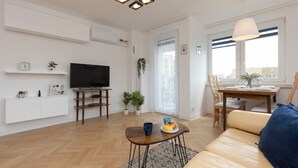 Una sala moderna con una televisión de pantalla plana, un comedor con una mesa y sillas de madera, y un sofá con un cojín azul.