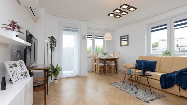 Una sala moderna con piso de madera, un sofá color beige, una mesa de comedor con sillas y un televisor sobre un mueble blanco.