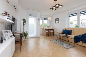 Een moderne woonkamer met een houten vloer, een beige bank, een eettafel met stoelen en een tv op een witte kast.