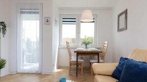 Una sala de estar con un sofá, una mesa de comedor con sillas, una mesa de centro con pasteles y una taza, y un ventanal con cortinas.