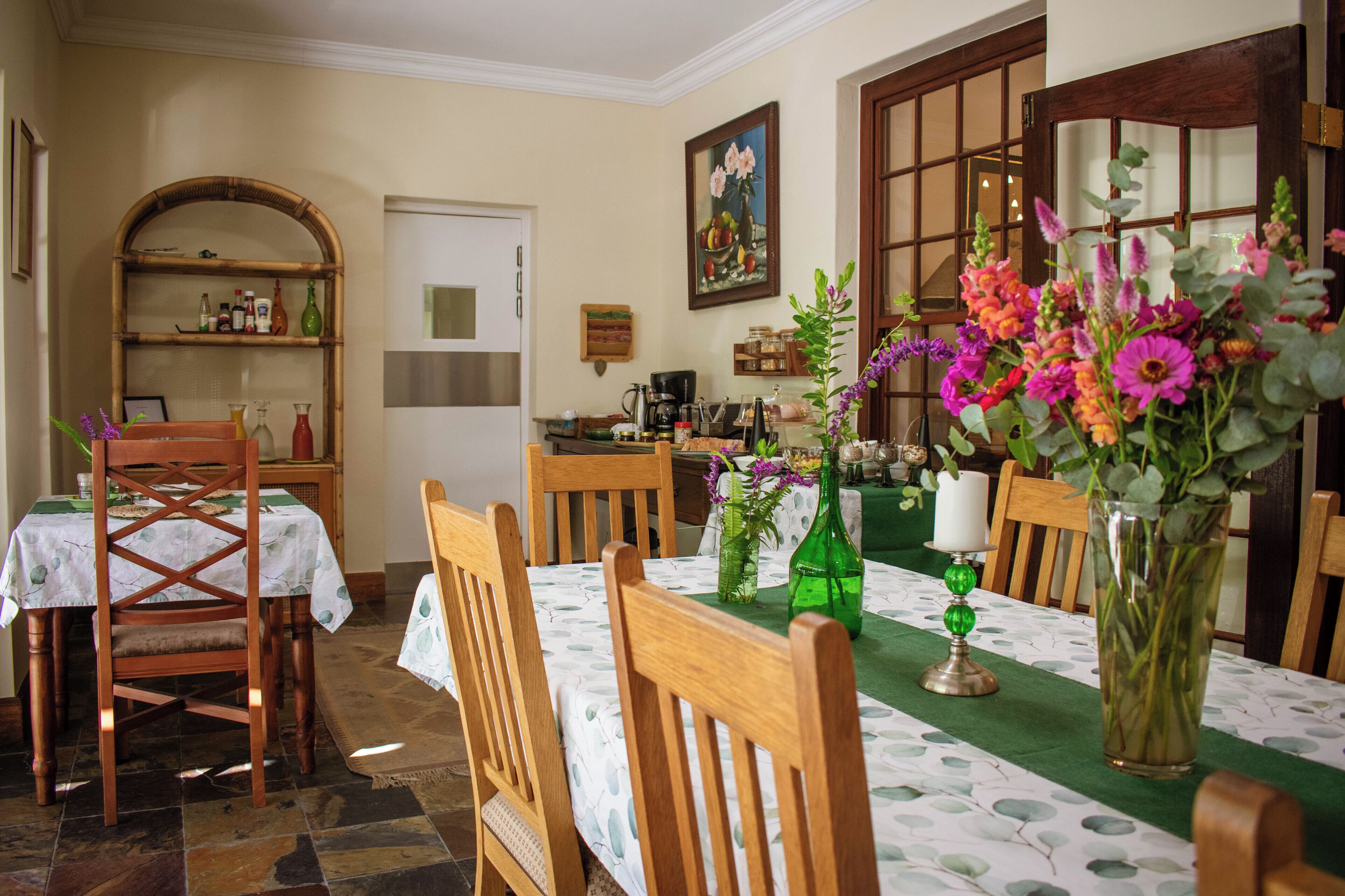 A dining area with a table set for four, surrounded by wooden chairs. A green vase with flowers is on the table, and a wooden shelf with various items is in the background.