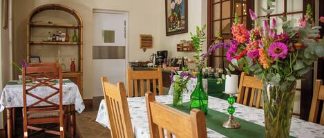 A dining area with a table set for four, surrounded by wooden chairs. A green vase with flowers is on the table, and a wooden shelf with various items is in the background.