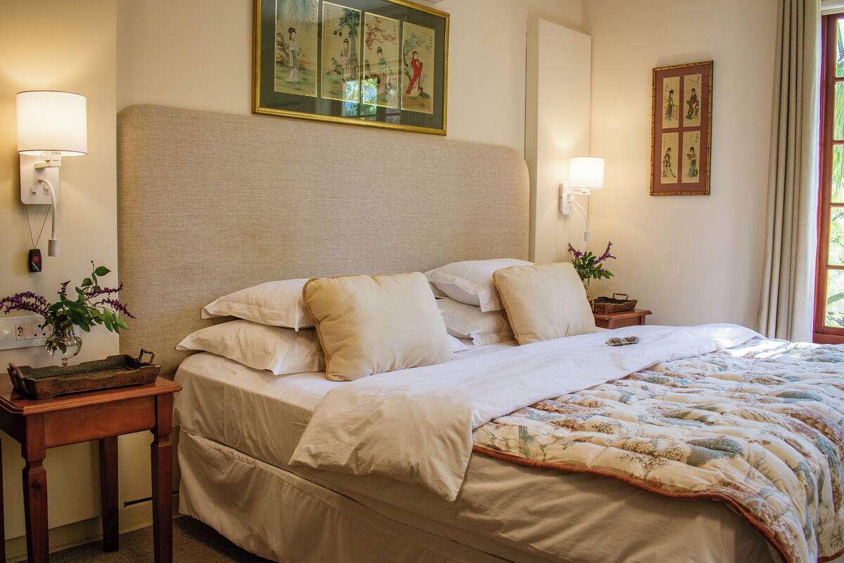 A neatly made bed with a floral comforter and multiple pillows, a bedside table with a lamp, and a wall-mounted painting.