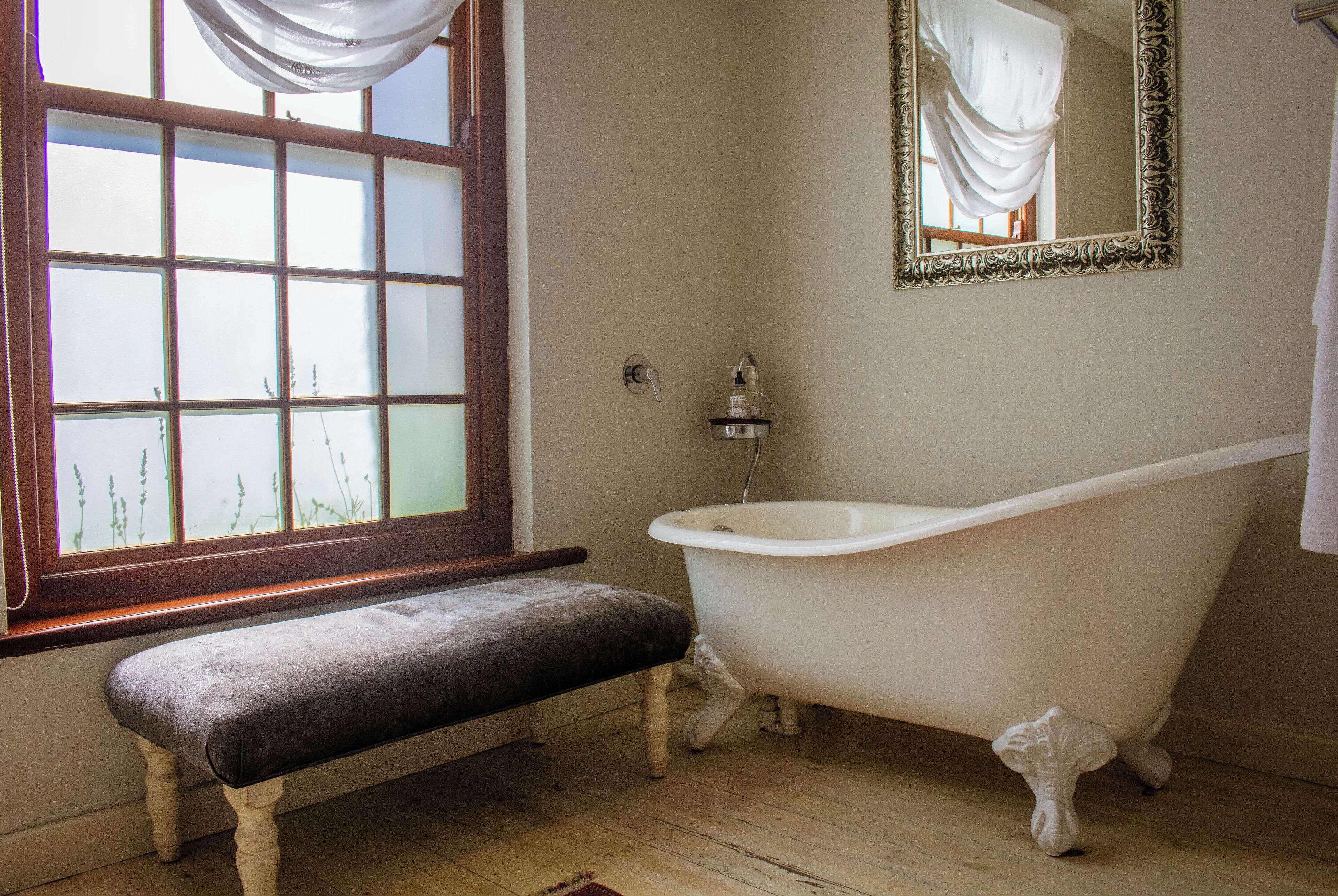 Un baño con una bañera tipo pata de león, un banco, una ventana con cortinas y un espejo con marco decorativo.